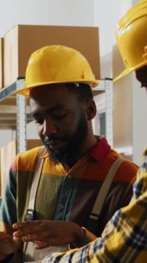 Vertical video: Male workers planning shipment with laptop in depot space, checking boxes with merchandise before doing distribution. Two men doing quality control in storage room, cargo stock.