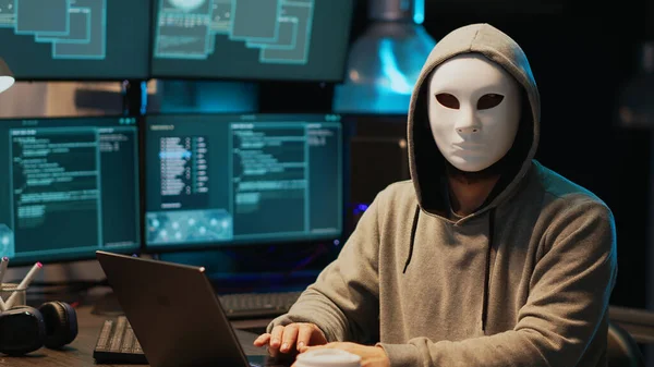 Hacker with anonymous mask breaking security system, installing virus to create computer malware and steal online data. Masked criminal scammer hacking network server late at night.