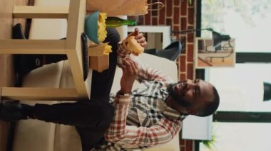 Dikey video: Neşeli bir adam oturma odasında hamburgerden bir ısırık alıyor, paket servis siparişinden fast food yiyor. Evde televizyon seyreden ve patates kızartması ve alkolle hamburger servis eden mutlu yetişkinler..