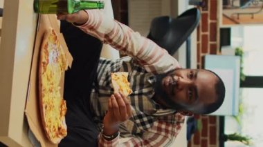 Dikey video: Genç adam kanepede pizza yiyor, dışarıdan yemek yiyor ve evde bira içiyor. Modern bir insan yemek ve alkol servisi yapar, TV 'de film izler..