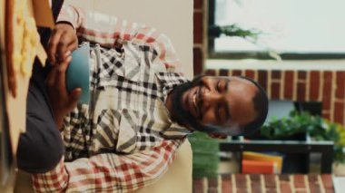 Dikey video: Afrikalı Amerikalı film bulmak için televizyon açıyor, kasede cips yiyor ve bira şişeleri içiyor. Genç, neşeli adam en sevdiği TV programını izliyor, çeşitli yemekler yiyor.