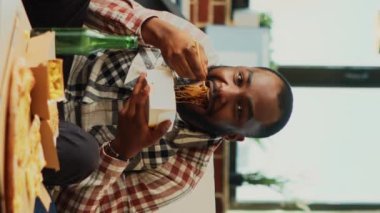 Dikey video: Afro-amerikan insanlar yemek çubuklarıyla erişte yiyorlar ve evde aksiyon dizileri izliyorlar. Genç ortaklar Asya yemeği yerken rahat hissediyorlar, film izliyorlar. Üçayak atışı.