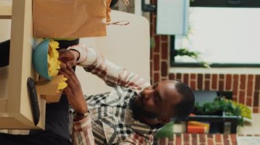Dikey video: Modern yetişkin akşam yemeğinde hamburger yiyor, hazır yemek yiyor ve televizyonda film izliyor. Genç adam hamburgerden zevk alıyor ve evde bira içiyor..
