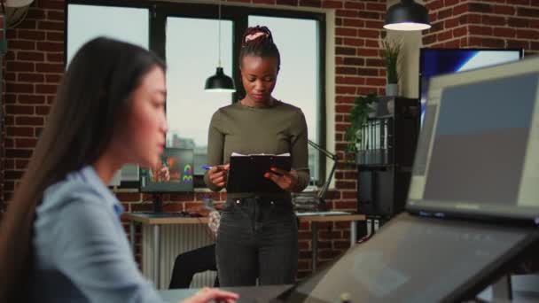 アフリカ系アメリカ人女性の従業員は デジタル代理店オフィスでドキュメントの仕事を読んで 笑顔アートデザイナー 3Dソフトウェアインターフェイスを使用する前にクリップボードファイルを見る女性 — ストック動画