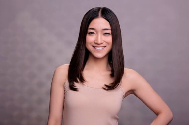 Çekici Asyalı kadın mutlu bir yüz ifadesi portresiyle eli kalçasında poz veriyor. Kendine güvenen genç ve doğal bir model bej bluz giyiyor ve gülümsüyor.