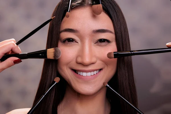 Makeup artist hands holding brushes and applying cosmetics on smiling cheerful asian model face. Make up professional arms using eye shadows and powder on young adult woman close up