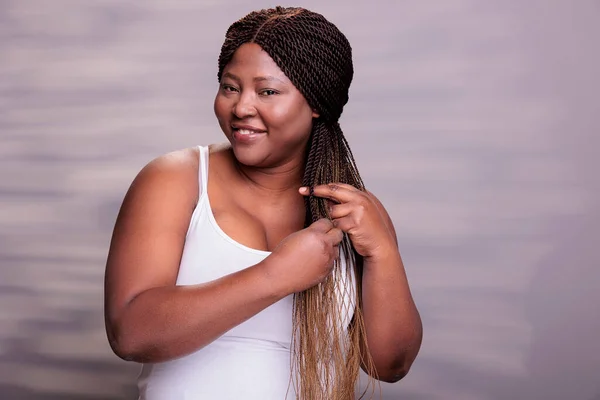 Plus size african american model making braids hairstyle and looking at camera. Beautiful happy body positive curvy woman smiling with cheerful facial expression braiding hair portrait