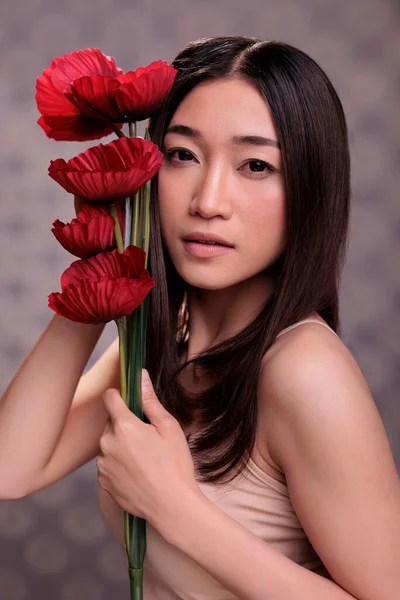 Beautiful asian woman posing for fashion portrait with red flowers bunch. Attractive beauty model using poppies bouquet to pose for natural cosmetology brand studio photoshoot