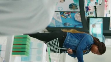 Vertical video: Female client taking boxes of pills and vitamins from drugstore, buying medicaments for healthcare. Young woman checking supplements and pharmaceutical products, medical store.