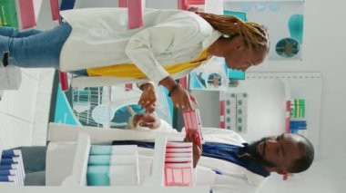 Vertical video: Male doctor recommending healthcare products to client, showing medicaments and supplements to cure disease. Young woman talking to specialist about treatment, buying box of pills.