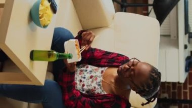 Vertical video: Cheerful girl serving noodles in delivery box, using chopsticks to eat asian food and watch action film on tv. Woman enjoying dinner from takeaway restaurant, having fun with