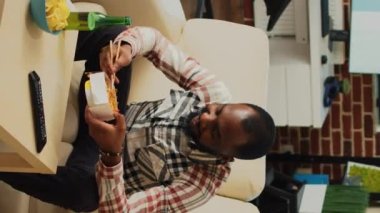 Vertical video: African american guy eating noodles in living room, sitting at television and watching movie. Young man serving asian food with chopsticks, ordering dinner from takeaway restaurant.