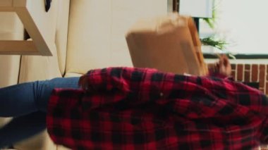 Vertical video: Smiling person ordering takeaway delivery meal at home, watching movie or tv show on television and eating fast food from takeout bag. Modern adult enjoying film on tv, drinking bottle