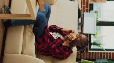 Vertical video: African american person taking bite of burger on couch, eating fast food from delivery a home. Young adult enjoying takeaway meal with fries and hamburger, watch movie on television.