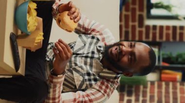 Dikey video: Afro-Amerikalı adam hamburger yiyip bira içerek eğleniyor, televizyonda komedi filmi izliyor. Yetişkin bir erkek fast food ve alkolün tadını çıkarıyor, oturma odasında televizyon seyrediyor..