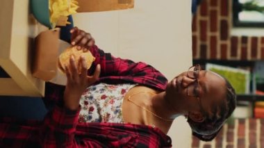 Dikey video: Patates kızartması ve birayla hamburger yiyen mutlu kadın, televizyonda aksiyon filmiyle eğleniyor. Hamburgerden ve atıştırmalıklardan hoşlanan gülümseyen kişi oturma odasında TV şovu seyrediyor..
