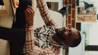 Dikey video: Afro-Amerikalı adam dışarıdan pizza yiyor, bira içiyor ve televizyonda kendini rahat hissediyor. Neşeli genç adam, aksiyon filmi ya da TV şovundan zevk alıyor.