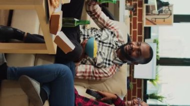 Dikey video: Afro-Amerikalı çift televizyon izlerken eğleniyor, hamburger, pizza ve nobles sipariş ediyorlar. Genç partnerler film ya da TV şovundan zevk alıyor, bira şişeleri içiyor..
