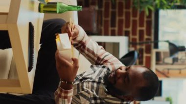 Vertical video: Male adult eating noodles with chopsticks at television, opening bottle of beer and feeling relaxed at home. Young man enjoying asian food delivery for dinner, watching film on tv