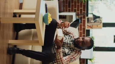 Vertical video: Modern guy eating noodles with chopsticks, holding box of asian food from takeaway order. Person enjoying tasty delicious meal from chinese takeout, watching action or comedy movie on