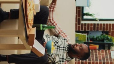 Dikey video: Bir şişe bira içen ve abur cubur yiyen genç, oturma odasındaki favori televizyon programının tadını çıkarıyor. Neşeli adam televizyonda filmle eğleniyor, paket burger, pizza ve...