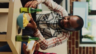 Vertical video: Happy man and woman eating asian meal at home, enjoying time together with takeaway food and alcohol bottles. Young couple using chopsticks to eat noodles at television. Tripot shot.