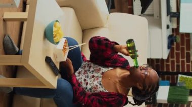 Dikey video: Afro-Amerikalı kız yemek çubuklarıyla erişte yiyor, bira servisi yapıyor ve kanepede en sevdiği TV programını izliyor. Yemek servisinden Asya yemeğinin keyfini çıkaran mutlu kadın, izle.