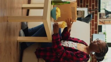 Vertical video: Young adult taking out hamburger and fries, eating fast food takeaway meal from delivery bag on couch. Woman enjoying dinner wth burger and snacks, watching comedy movie on television.