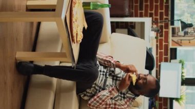 Vertical video: Cheerful adult taking bite of pizza at home, eating takeaway food delivery and drinking alcohol. Young relaxed man having fun watching action movie on television, eating snacks.