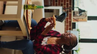 Dikey video: Afro-Amerikan kadın televizyon izliyor ve pizza yiyor, oturma odasında komedi filmine gülüyor. Genç insan fast food servisi yapıyor ve en sevdiği TV programını izliyor..