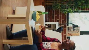 Vertical video: Modern person using chopsticks to eat noodles, drinking bottle of beer while she watches tv show at home. Young adult enjoying asian food from delivery order bag, watching movie.