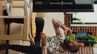 Dikey video: Mutlu adam pizza yiyor ve evde bira içiyor, dışarıdan yemek yiyor ve televizyon seyrediyor. Akşam yemeği ve alkol servisi yapan genç adam, oturma odasında film izliyor..