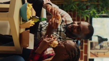Vertical video: African american people eating burgers and fries, watching movie on television and laughing together. Happy couple feeling relaxed with fast food takeaway meal, drinking beer bottles.