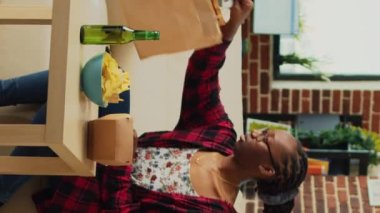 Dikey video: Fast food servisinden hamburger yiyen neşeli insan, o TV 'de film veya komedi programı izlerken hamburger ve patates kızartmasıyla yemeğin tadını çıkarıyor. Eve paket servis yapan bir kadın..