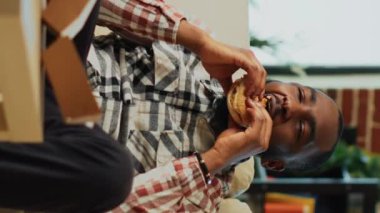 Dikey video: Afro-amerikalı bir adam hamburgeri patates kızartmasıyla ısırıyor, eve servis edilen yemekleri yiyor. Mutlu insan fast food yiyip bir şişe bira içip rahatlıyor.