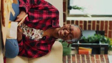 Vertical video: Happy female adult laughing at comedy film on tv, relaxing on couch with beer and snacks. Young woman havving fun at home with movie on television, takeaway delivery. Tripod shot.