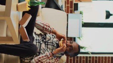 Vertical video: African american person eating burger and fries on sofa, having dinner with fast food and bottle of alcohol at home. Young man feeling happy watching action movie or show, having fun.