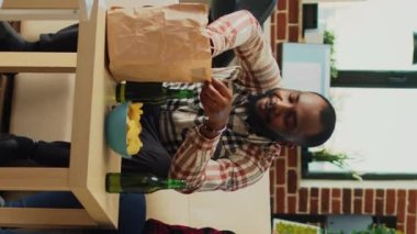 Vertical video: African american couple eating burgers with fries at home, having fun watching movie on tv and drinking beer. Boyfriend and girlfriend serving fast food meal from takeaway restaurant.