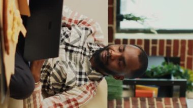Vertical video: Smiling man using laptop in living room and watching tv, feeling relaxed with multiple delivery meals and beer. Young guy browsing internet on pc, sitting in front of television