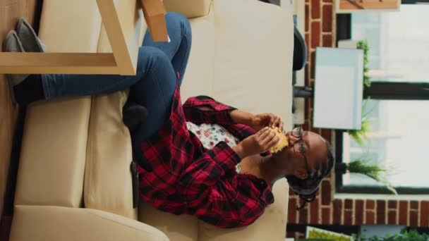 Vertikales Video Afroamerikaner Beißt Burger Auf Couch Und Isst Fast — Stockvideo