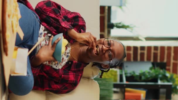 Vertikales Video Afroamerikanerin Isst Schüssel Chips Auf Sofa Und Lacht — Stockvideo