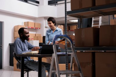 Storehouse employees having fun at job while preparing clients packages putting order in carton box, working in warehouse delivery department. Diverse team checking cargo stock on laptop computer