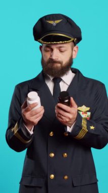 Vertical video: Male aviator looking at bottles of pills in studio, feeling unsure about medicaments and medical treatment. Young adult flying pilot holding jars of medicine and supplements, aviation