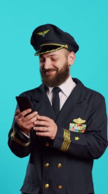 Dikey video: Uçuş ekibi kaptanı, stüdyoda internet üzerinden akıllı telefon kullanarak sosyal medya uygulamalarını tarıyor. Mavi arkaplan üzerinde cep telefonu ve tarayıcıyla oynayan genç uçak pilotu.