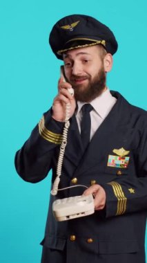 Vertical video: Commercial pilot talking on landline phone call in studio, answering office telephone with cord for remote conversation. Young smiling airliner in uniform chatting on phone line.