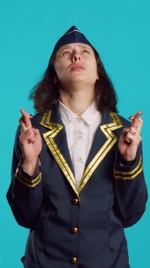 Vertical video: Positive air hostess standing with fingers crossed on camera, asking to fulfill wish over blue backdrop. Female aircrew member praying for luck and fortune, acting hopeful and wishful.
