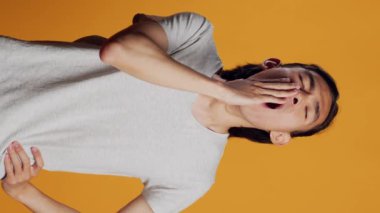 Vertical video: Drained person yawning and being tired in studio, falling asleep and feeling worn out. Exhausted young man being sleepy and carefree over orange background, having confidence on camera