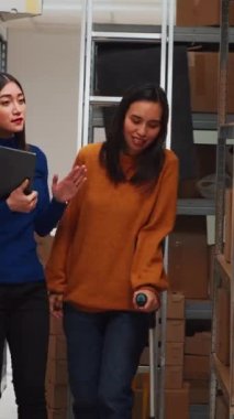 Vertical video: Young person in crutches checking boxes on shelves, looking at products stock in storehouse space and working with gadget. Team of people planning distribution for business plan