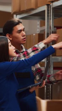 Vertical video: Team of workers analyzing cardboard boxes on shelves, checking merchandise stock in warehouse. Man and woman using laptop to work on products shipment, small business development.