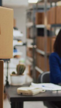 Vertical video: Female entrepreneur working on supply chain development with products stock in packages, planning distribution in warehouse. Young woman using laptop and checking quality of goods.